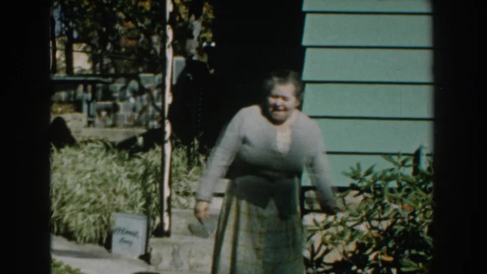 The woman leans forward near the front shrubbery, framed by porch steps, a dark doorway, and green siding in the 1959 Michigan clip.