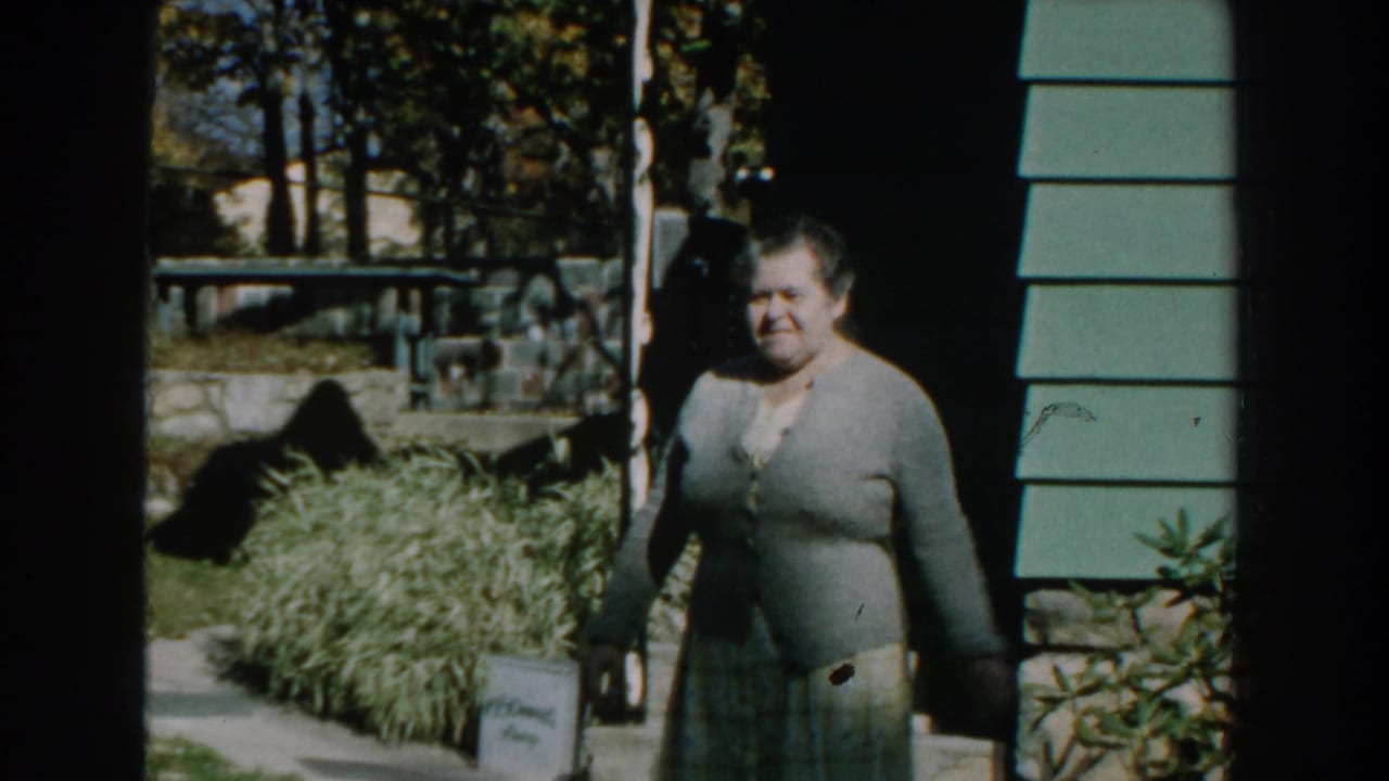 Woman in a gray cardigan stands near a house entrance beside a lawn and trees.
