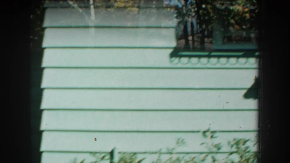 A close, off-center home-movie frame shows painted house siding with partial exterior details and clipped yard elements.