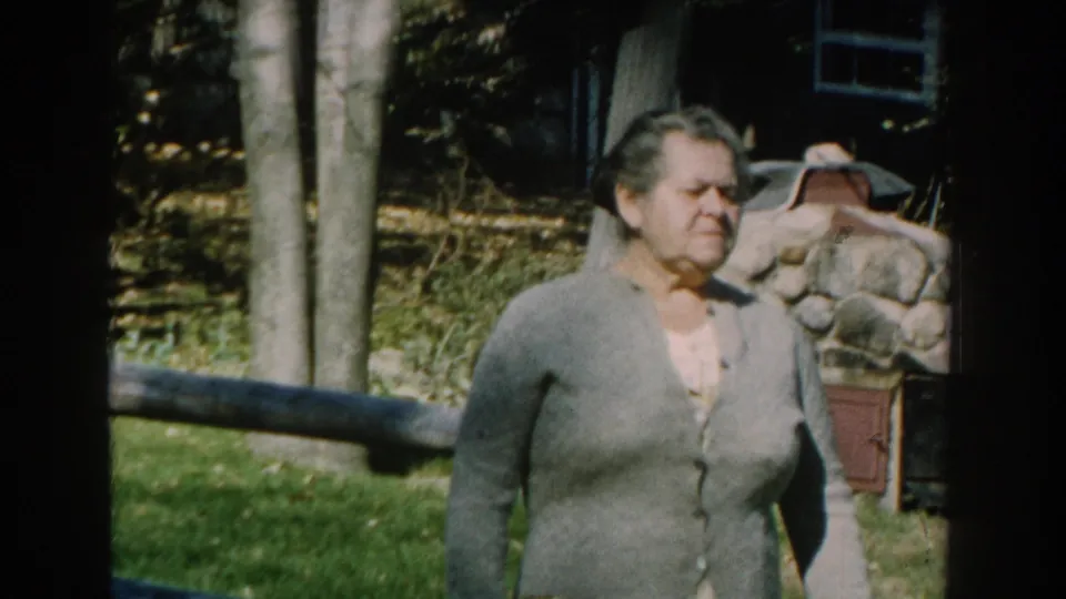 An adult woman in a gray cardigan and skirt walks through a Michigan garden area, with trees, grass, fencing, and stonework behind her.