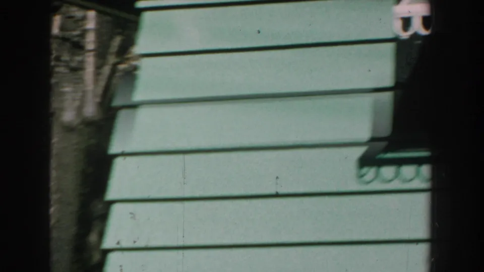 1959 Michigan home-movie frame of light green horizontal siding, deep shadow, and a narrow strip of stone wall.