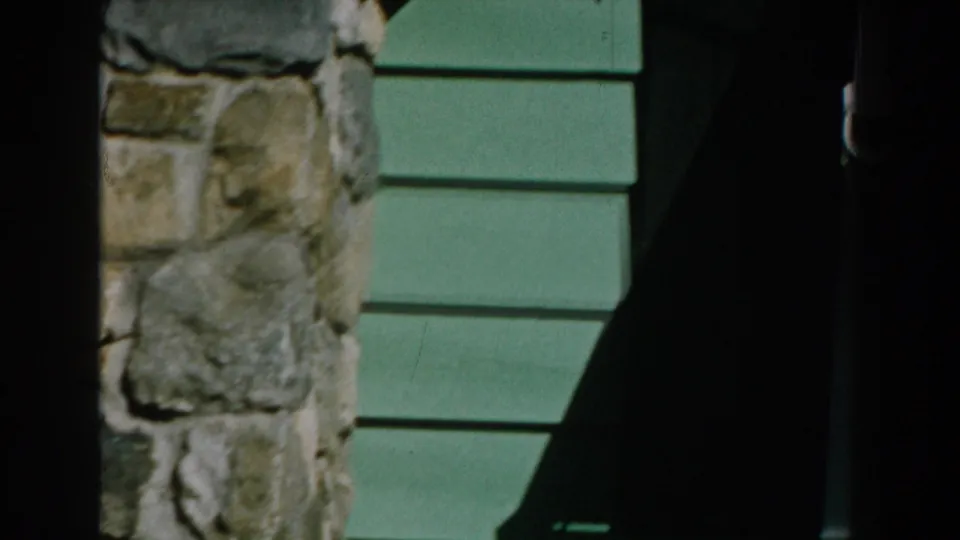 1959 Michigan frame with a stone wall section meeting green horizontal siding under strong shadow.