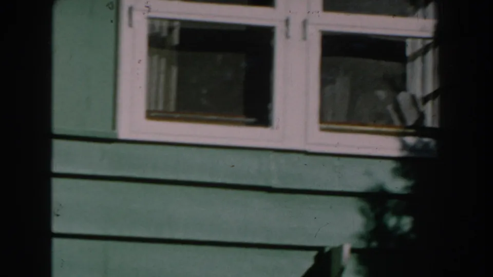 1959 Michigan 8mm frame showing green horizontal siding, a white-trimmed window, and shaded foreground obstruction.