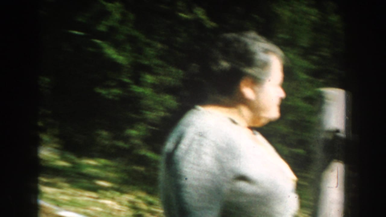 Older woman in a light ash-colored sweater walks through a leafy Michigan yard in 1959 home-movie footage.