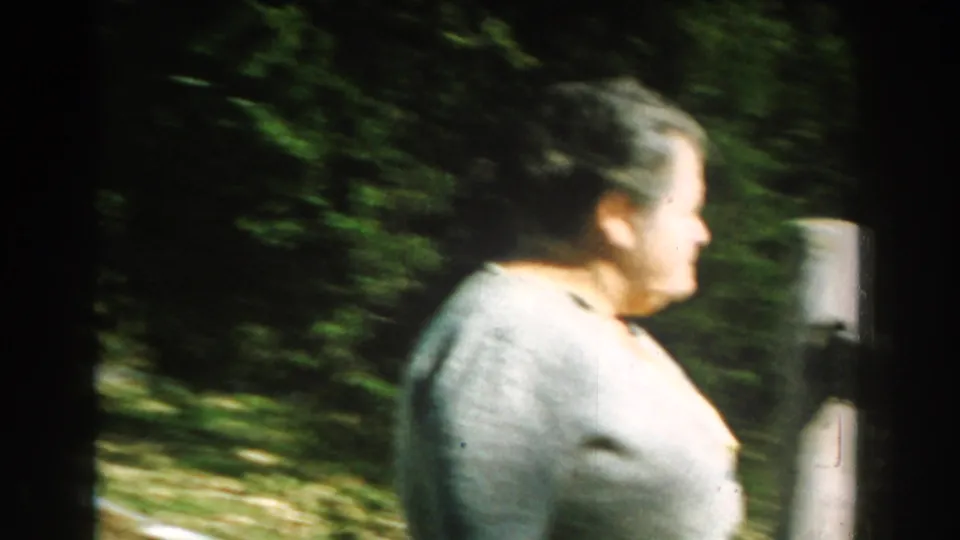 Older woman in a light ash-colored sweater walks through a leafy Michigan yard in 1959 home-movie footage.