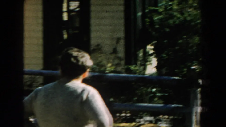 The woman is seen from behind near a wooden fence, with house siding and foliage in the background.