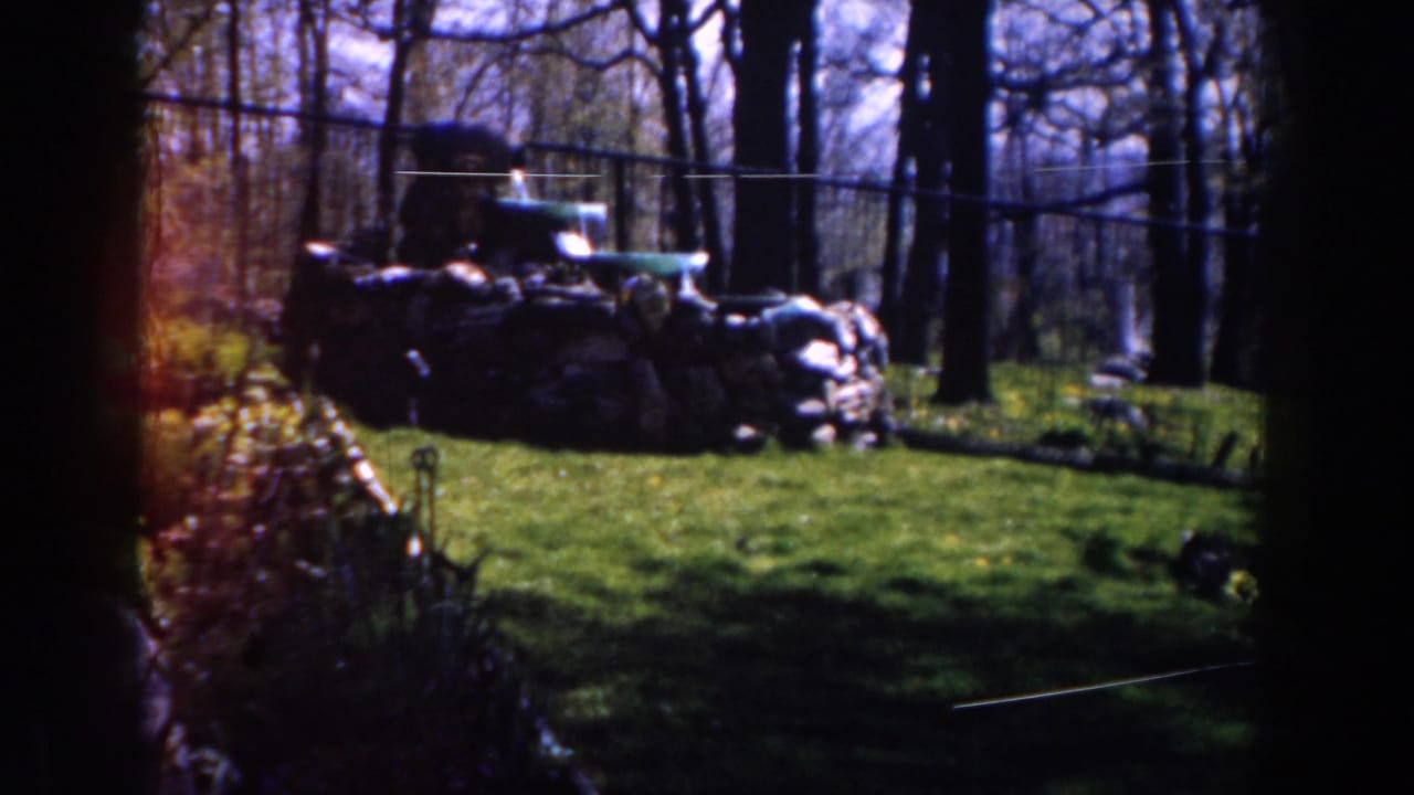 1959 Michigan 8mm frame showing a shaded wooded clearing with heaped sacks, a low wall, and a green vehicle in the background.
