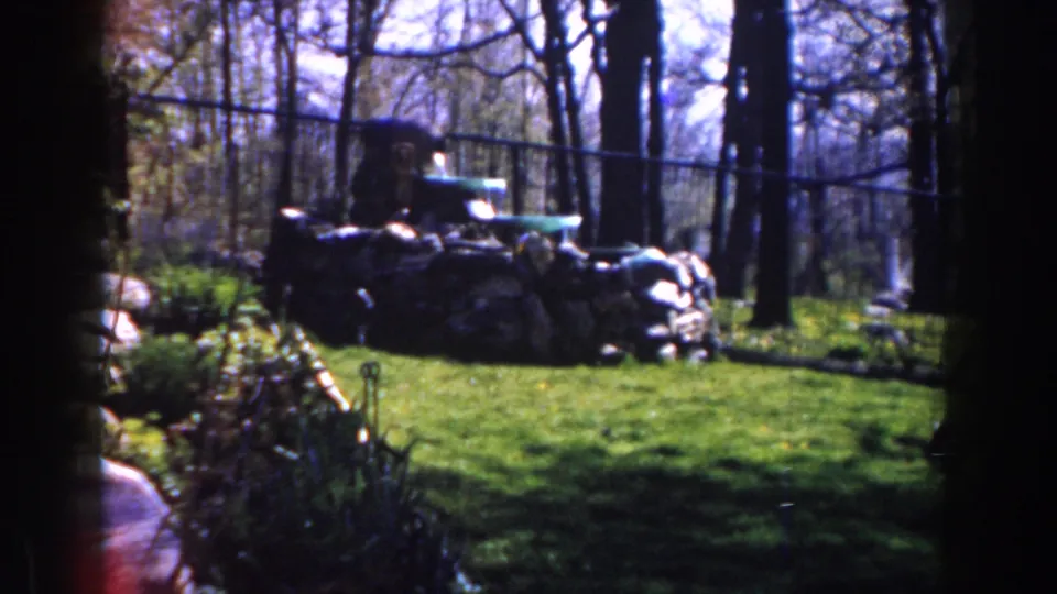 1959 Michigan home-movie frame with a green vehicle behind heaped sacks or sandbags in a sunlit forest clearing.