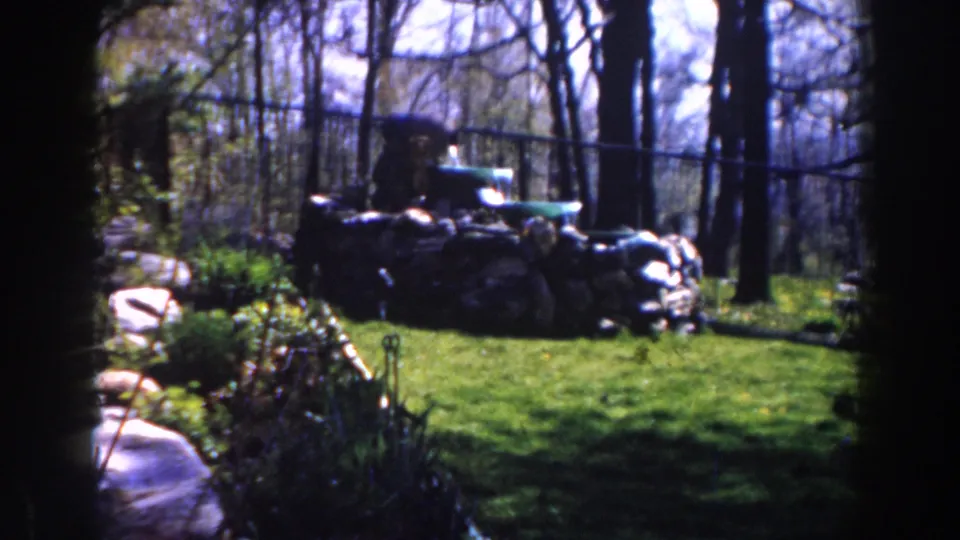 Soft 1959 8mm frame from Michigan showing piled sacks, a green vehicle, trees, and garden-like foreground foliage.