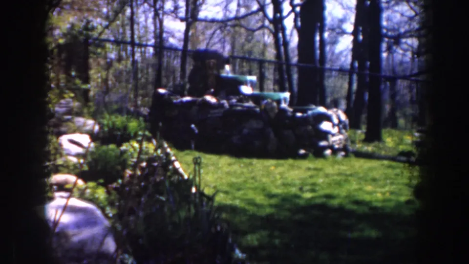 1959 Michigan frame showing a wooded camp-like clearing with heaped sacks, a low stone wall, and a green vehicle beyond.