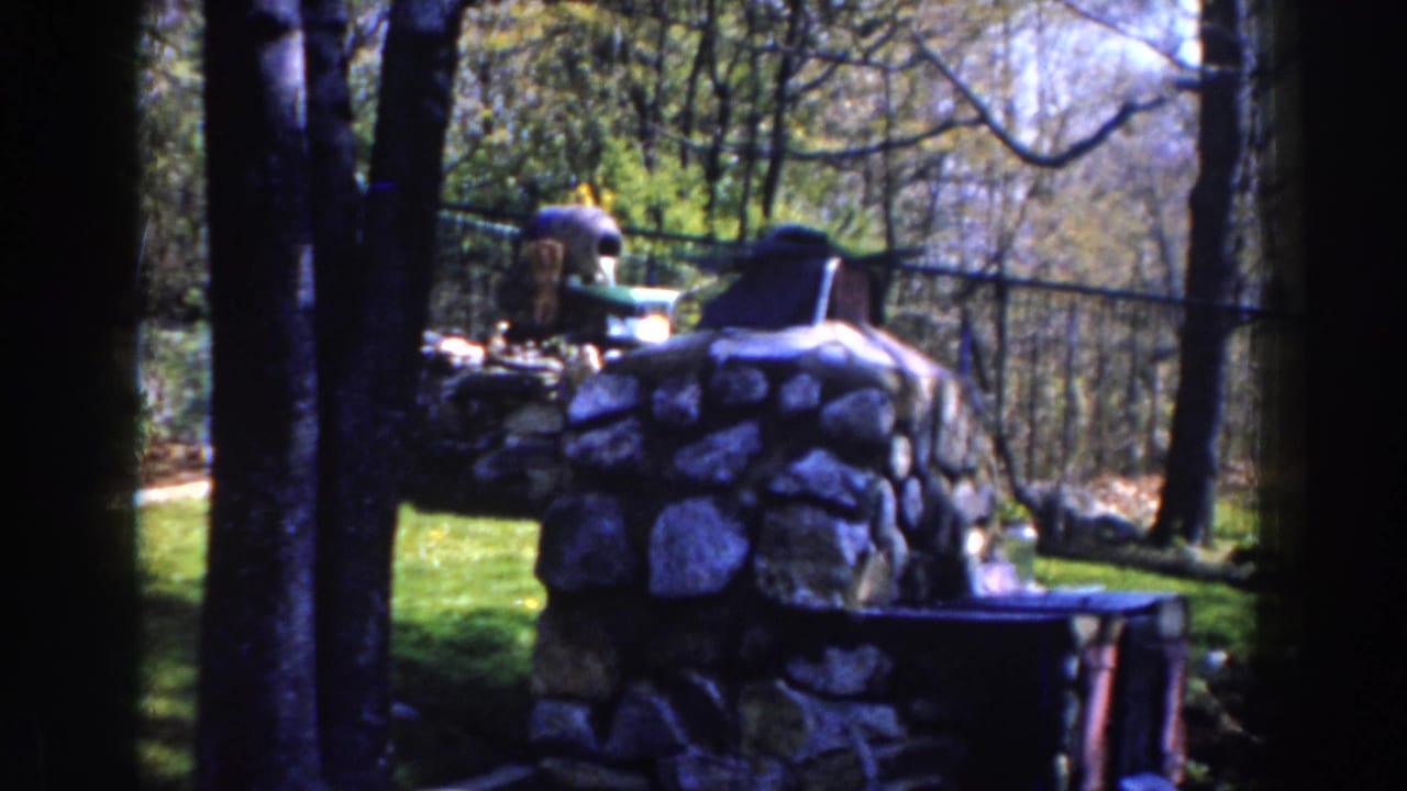 1959 Michigan 8mm frame showing a rough stone memorial near a dark metal fence, trees, grass, and a small wooden feature.