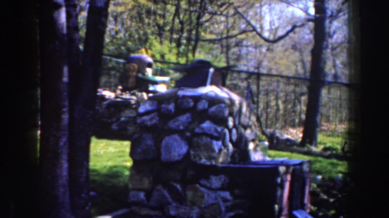 1959 Michigan frame of a fenced outdoor memorial area with a stone marker, lawn, bare branches, and bright spring light.