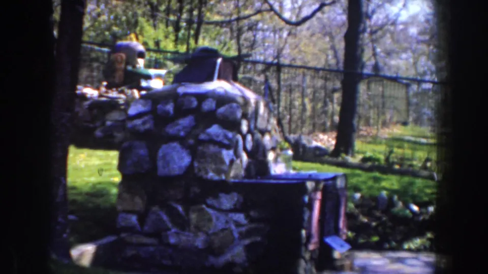 1959 Michigan 8mm frame with the stone memorial larger in view, a dark fence behind it, trees overhead, and sunlit grass beyond.
