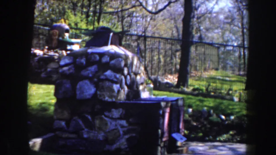1959 Michigan 8mm frame showing a rustic stone outdoor fireplace beside a grassy, tree-lined backyard with fencing behind it.