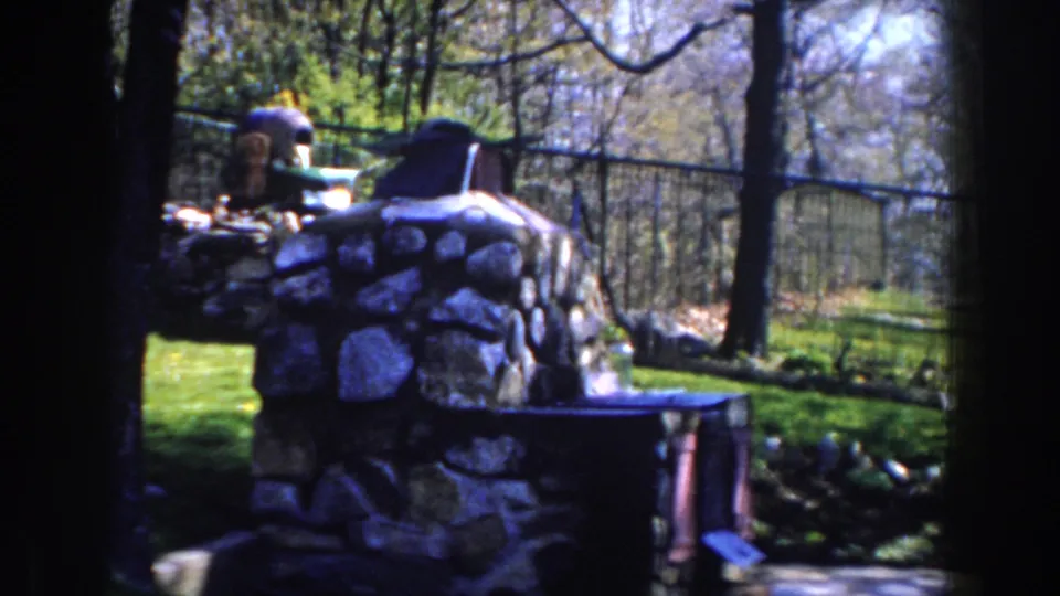 1959 Michigan backyard view with a stone fireplace, green lawn, trees, and wire fencing in soft 8mm color.