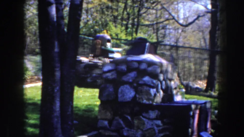 1959 Michigan home-movie frame of a stone backyard fireplace seen through nearby trees, with lawn and fencing beyond.