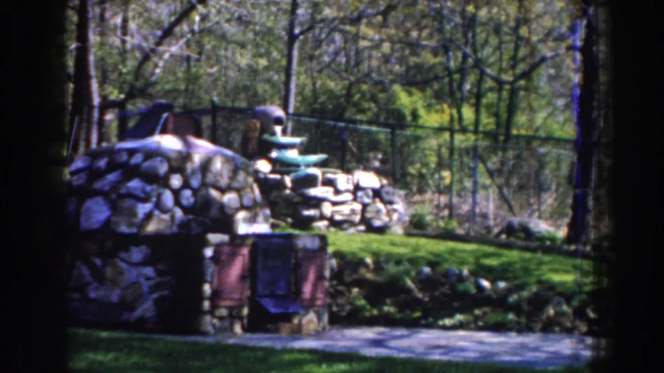 Wider 1959 Michigan 8mm view of the stone outdoor fireplace set into a grassy backyard with trees and fencing behind it.