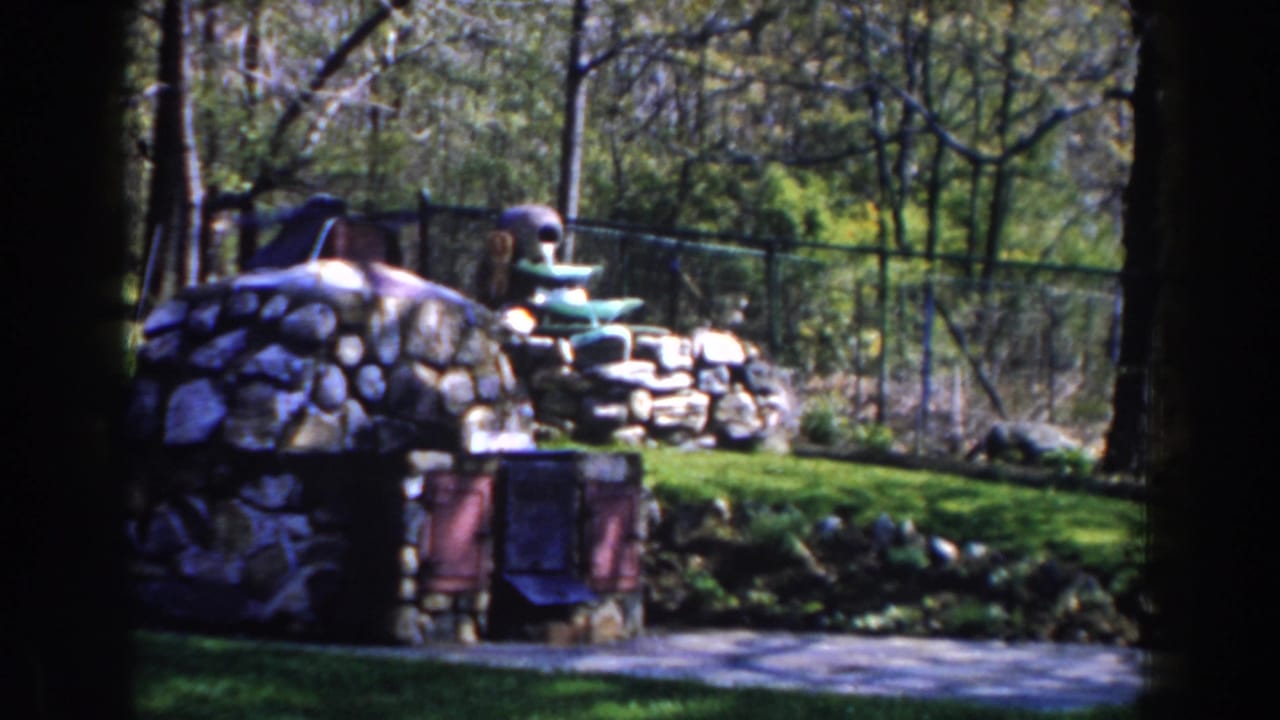 Stone fountain and low stonework in a grassy garden with trees and a chain-link fence.