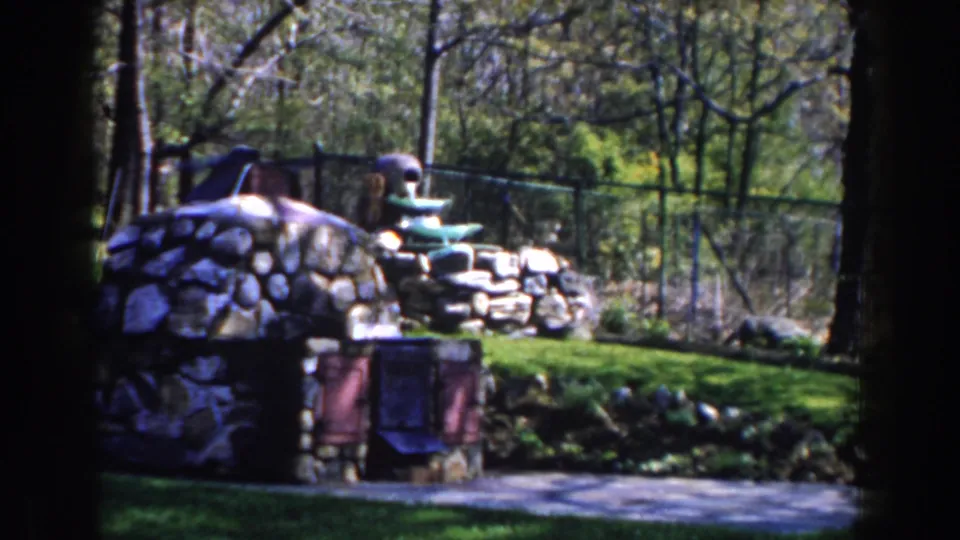 1959 Michigan 8mm frame showing a tiered stone fountain in a green garden bordered by trees and chain-link fencing.