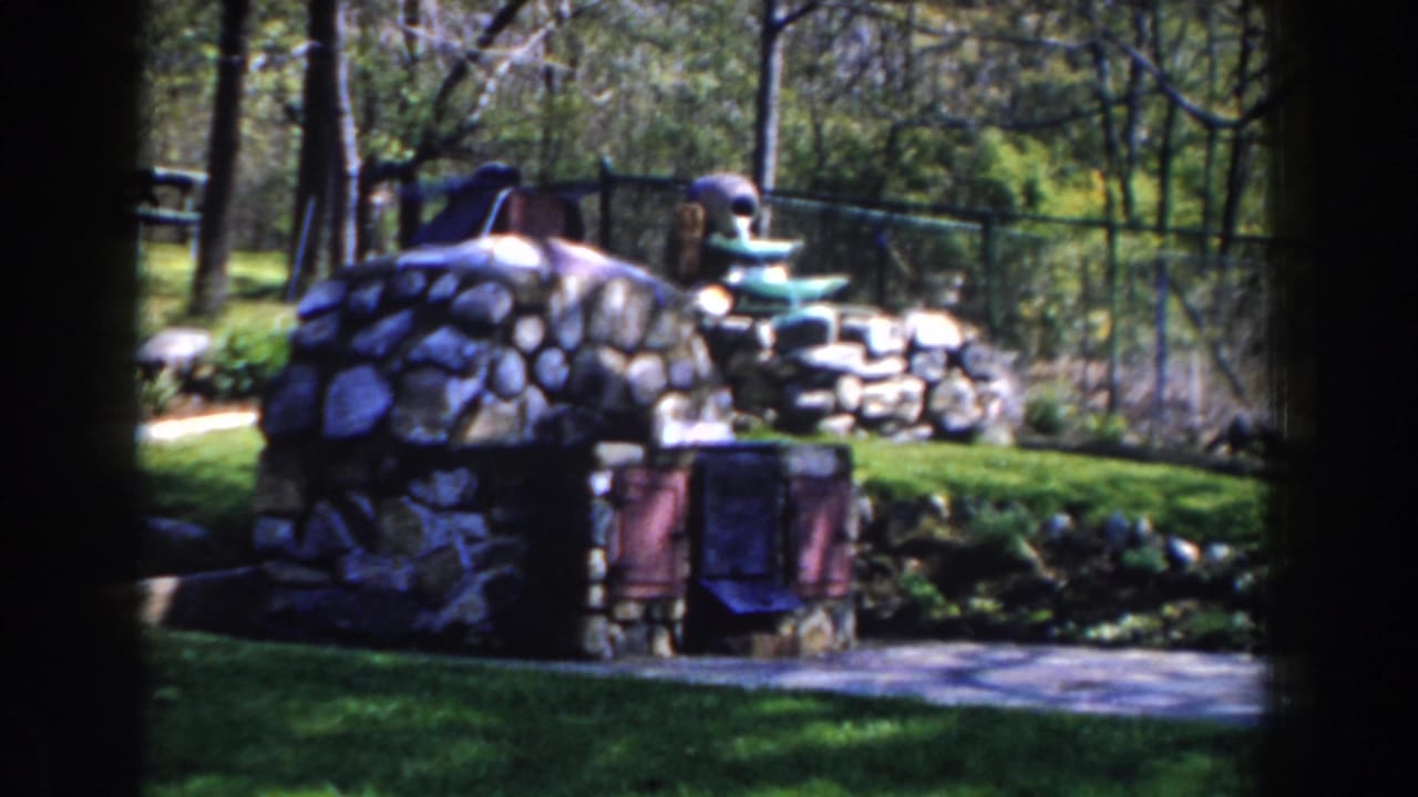 Garden fountain beside stonework, grass, trees, and a fence in bright sunlight.