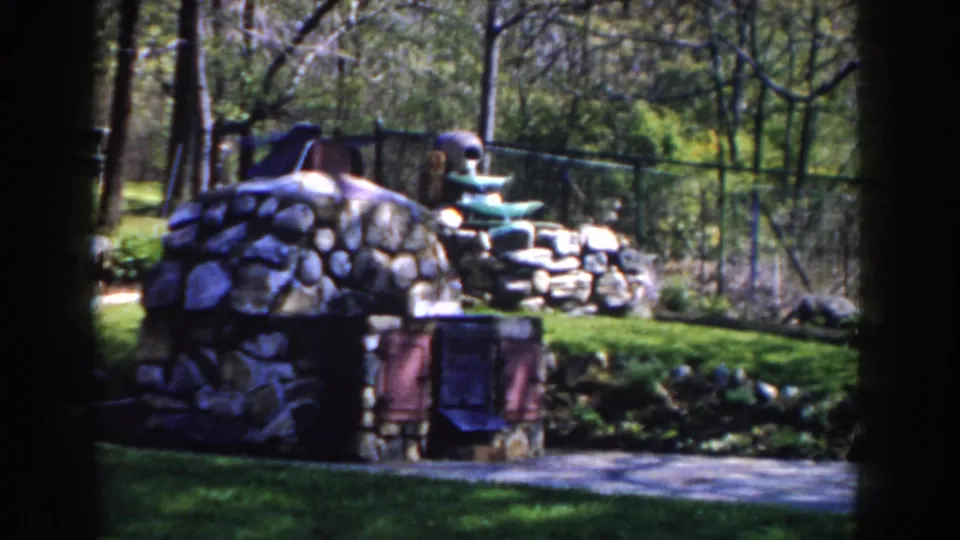 1959 Michigan 8mm view of a tiered stone fountain, low wall, grass, trees, and fence, partly framed by shadow.