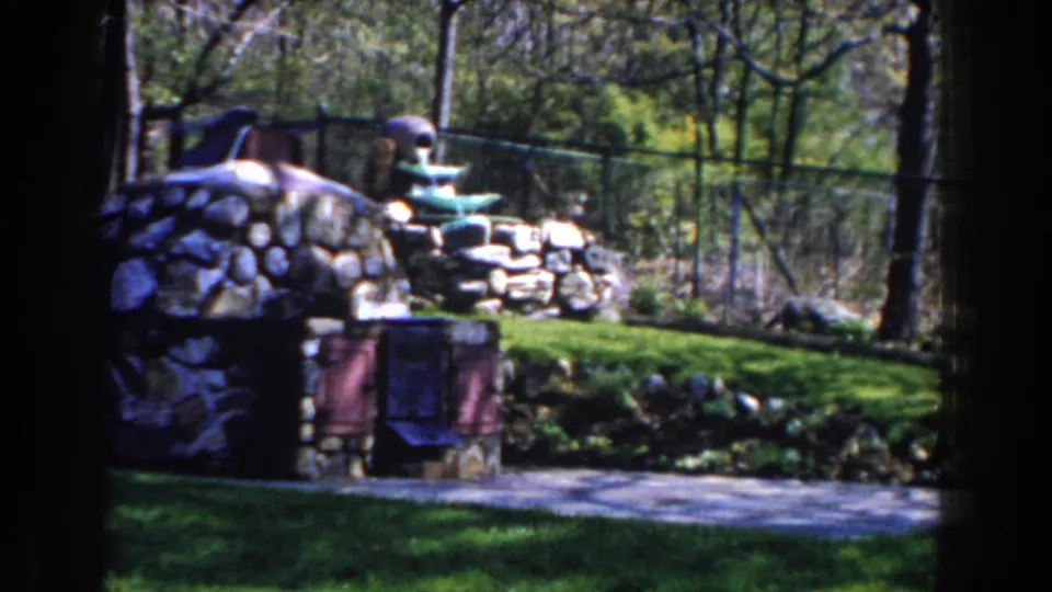1959 Michigan 8mm frame showing the garden fountain from a similar angle with stonework, lawn, trees, and fence visible.