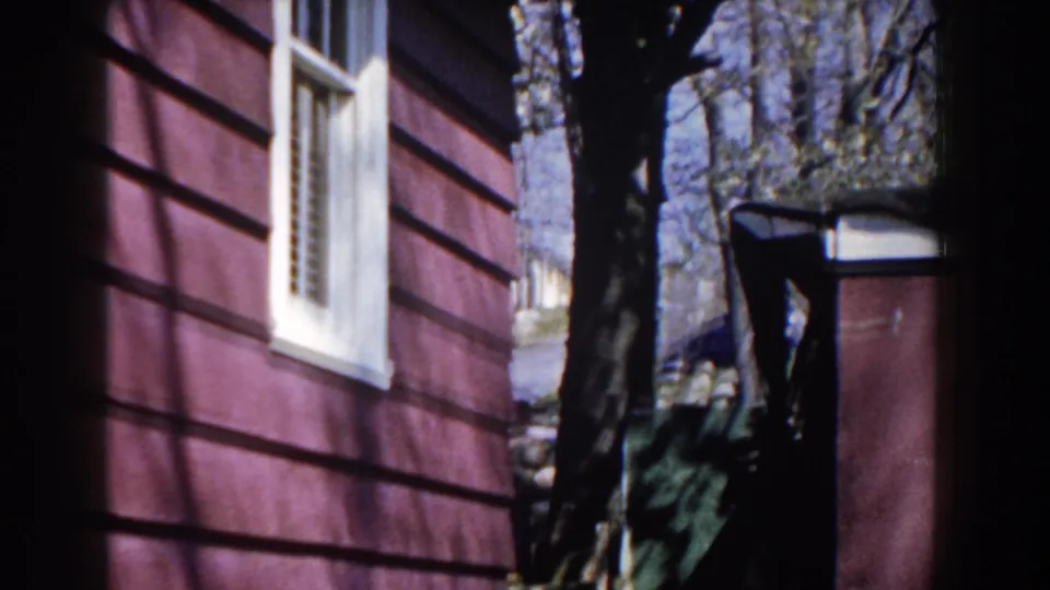 Red-sided Michigan house exterior with a white-trimmed window, tree trunks, and yard structures in 1959 home-movie footage.
