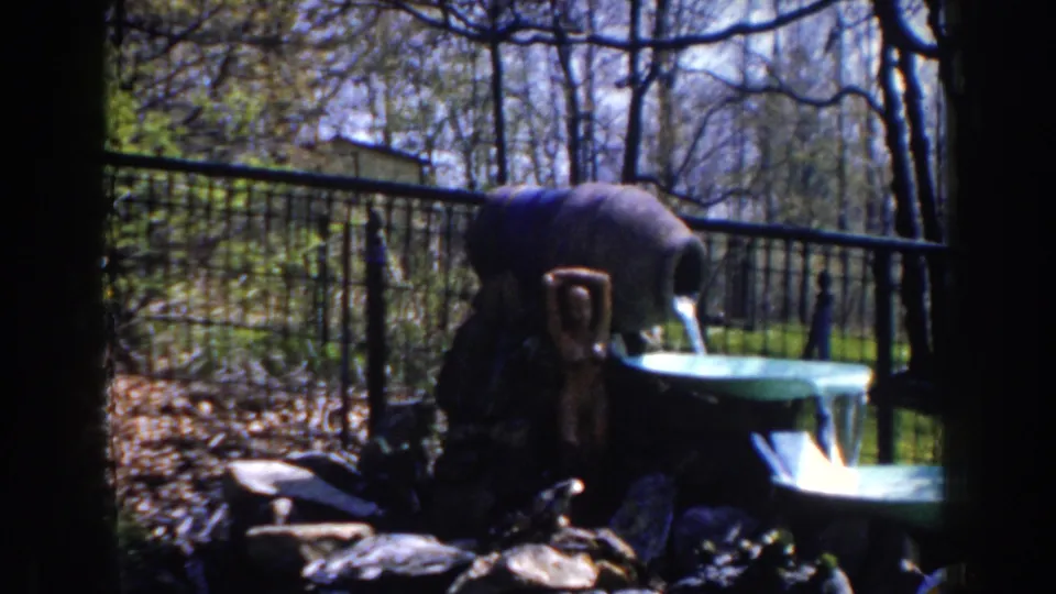 1959 Michigan 8mm frame showing a backyard-style fountain with a ceramic pitcher, green basin, rocks, fence, trees, and lawn.