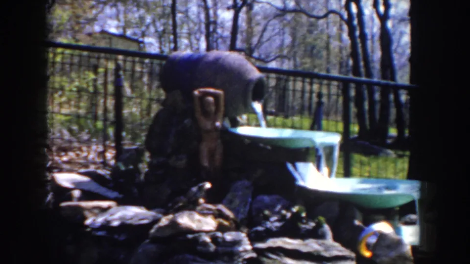 1959 Michigan home-movie frame of a small outdoor fountain, with a pitcher pouring into green basins beside rocks and a black fence.