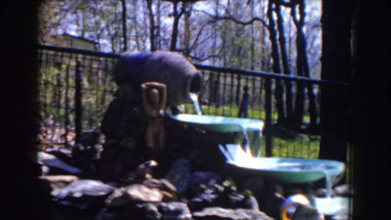 1959 Michigan 8mm frame showing a green tiered garden fountain, flowing water, dark rocks, metal fencing, grass, and bare trees.