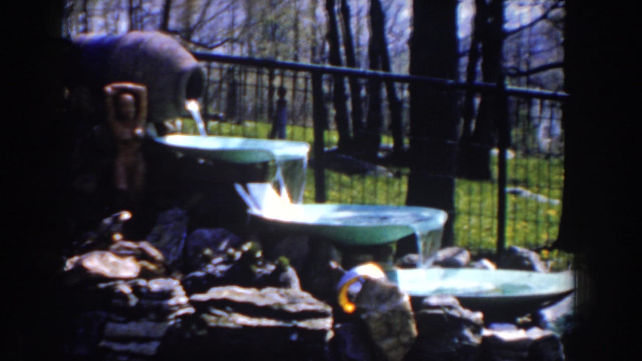 Tiered green fountain and rocks in front of fencing, grass, and leafless trees.
