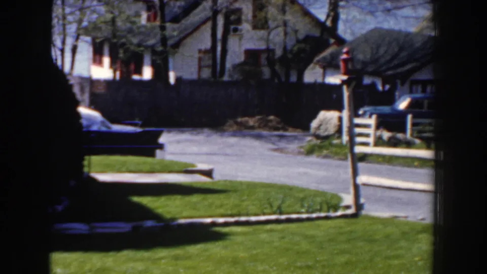 Late-1950s Michigan residential exterior showing a green lawn, driveway, fence, neighboring house, trees, and a partly visible parked car.