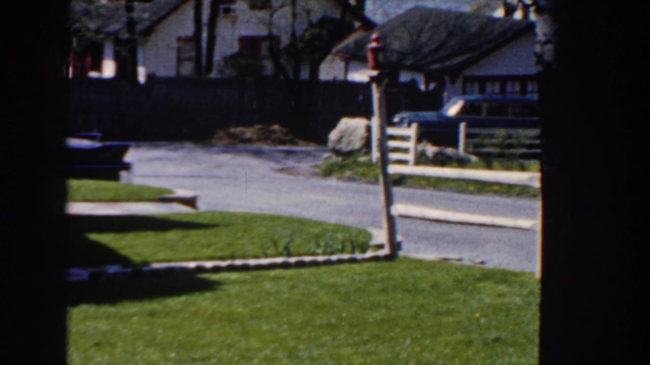 Quiet residential street with green lawn, curb, houses, a fence, and a parked dark car.