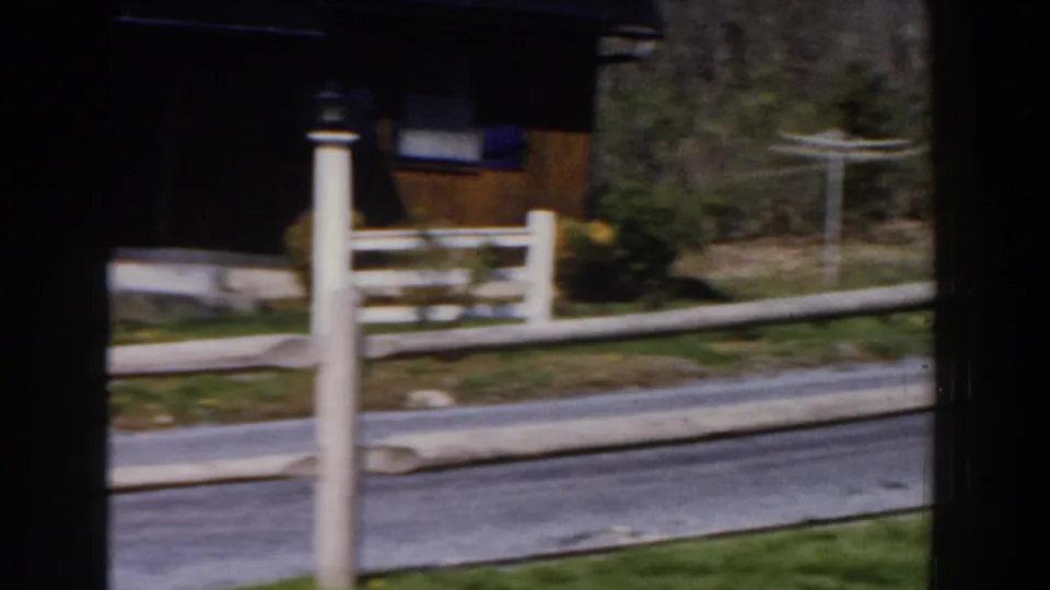 Home-movie pan in a 1959 Michigan neighborhood, showing fence posts, lawn, street edge, trees, and part of a house exterior.