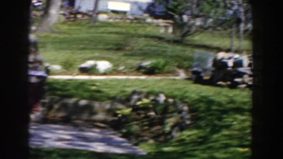 1959 Michigan home-movie frame showing a green suburban backyard, stone landscaping, trees and a house in the background.