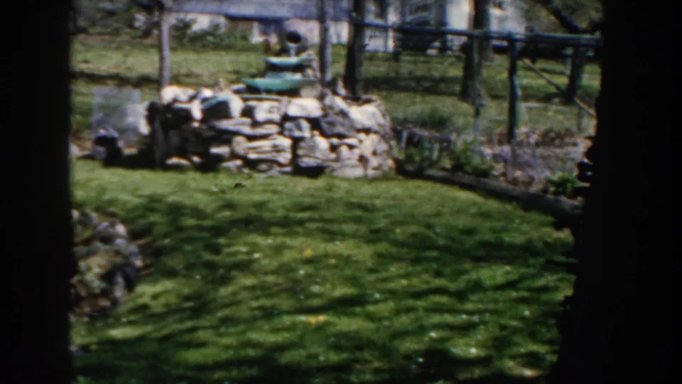 1959 Michigan home-movie still showing the backyard fountain and stone wall from a slightly closer view across the lawn.