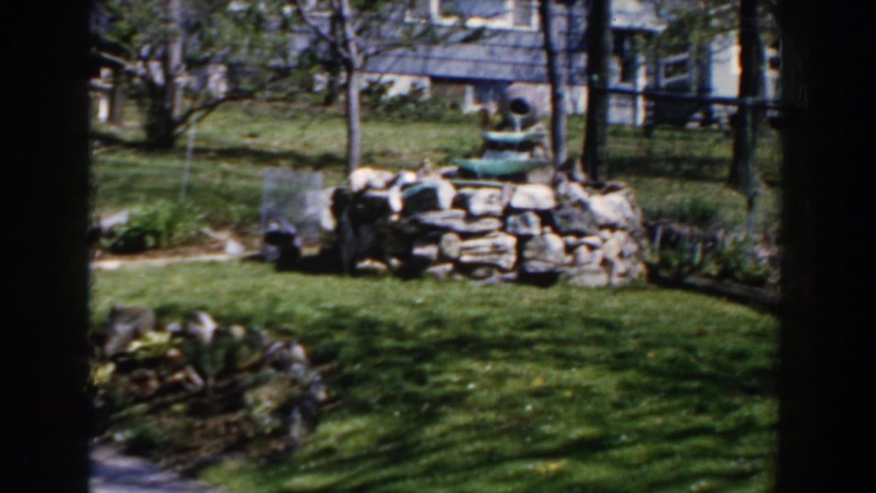 Backyard lawn with a low stone fountain, garden beds, trees, and a house in the background.