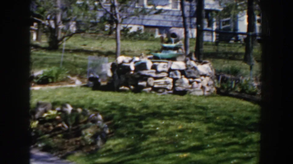 1959 Michigan home-movie frame showing a sunny backyard with grass, stone fountain, young trees, and a house beyond.
