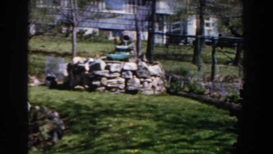 1959 Michigan backyard view with a tiered stone fountain, green lawn, trees, and house exterior in soft 8mm color.