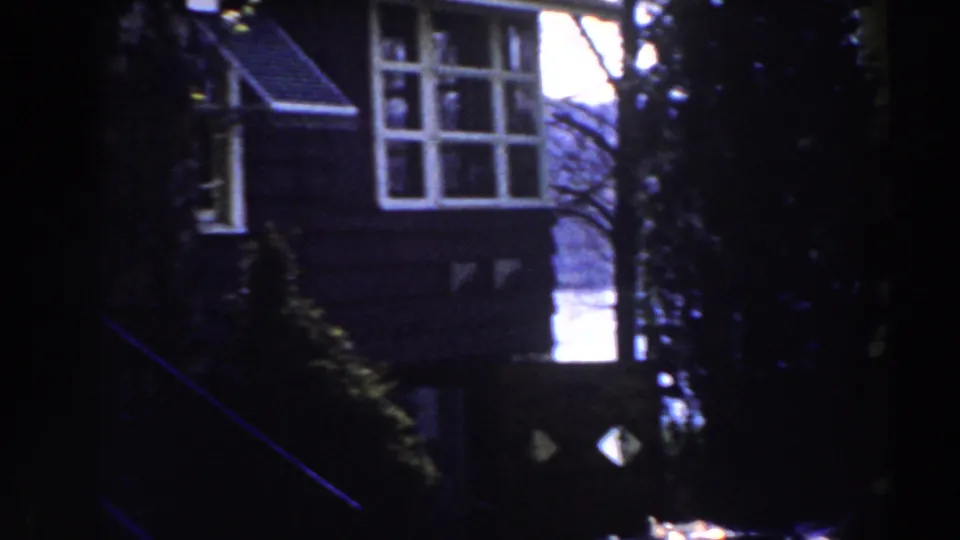 1959 Michigan home-movie frame showing a house corner with multi-pane windows, a striped awning, shrubs, and bright sky behind trees.