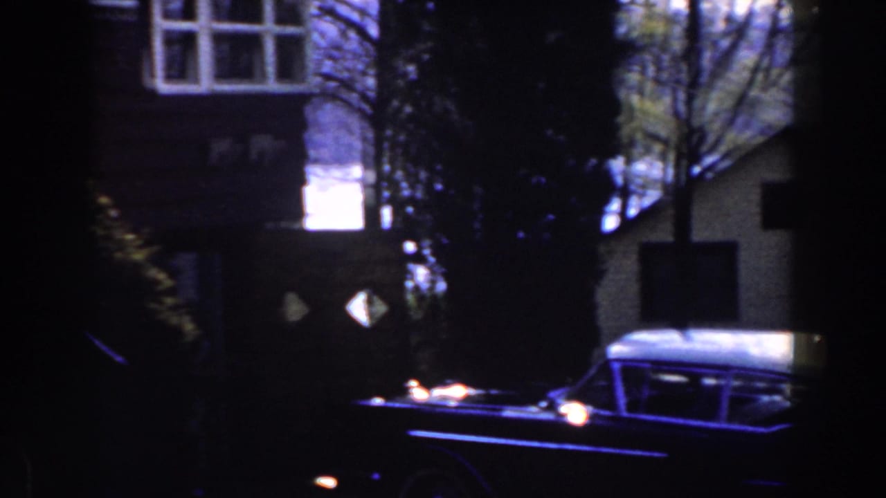 1959 Michigan home-movie frame showing a dark foreground edge, a large finned car, houses, trees, and water in the background.