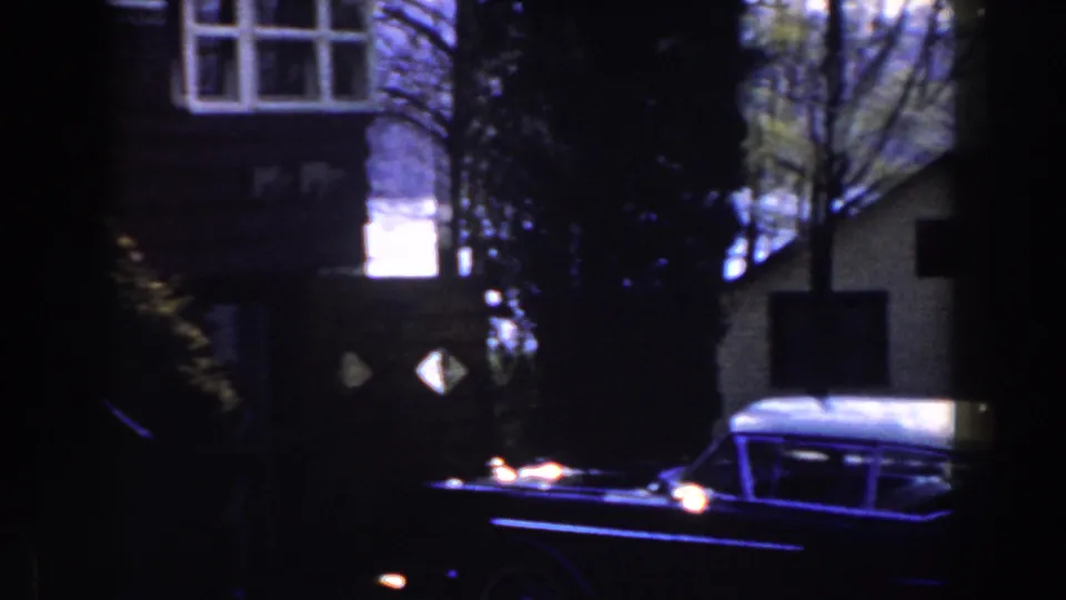 1959 Michigan home-movie frame showing a dark foreground edge, a large finned car, houses, trees, and water in the background.