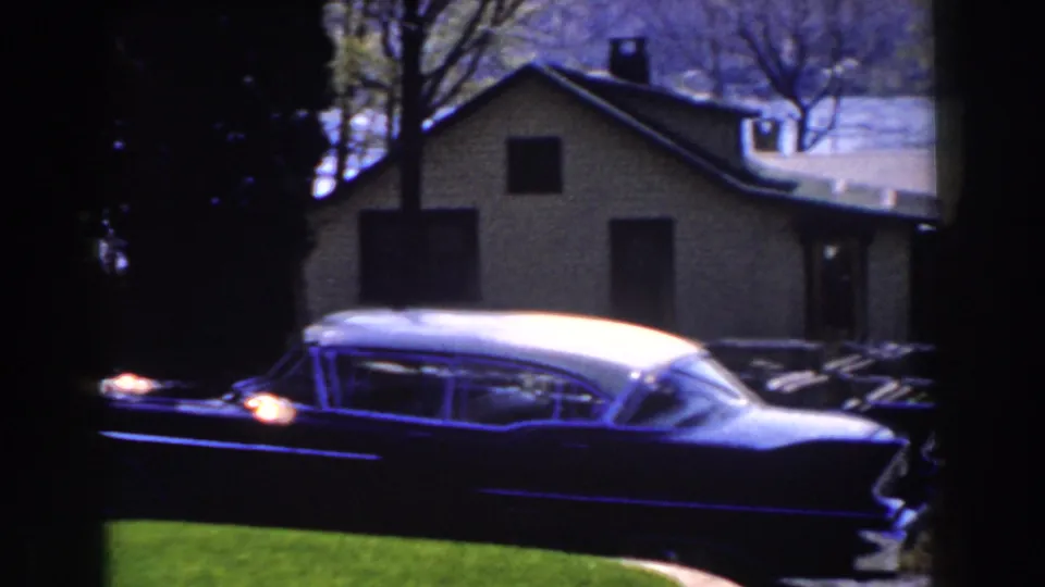 A late-1950s two-tone car with a white roof is parked near grass, a driveway, bare trees, and a modest Michigan house.