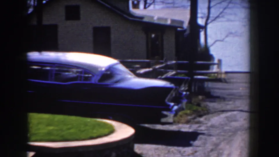The camera catches the rear side of the finned car by a curved lawn edge, driveway pavement, houses, and trees in Michigan home-movie footage.