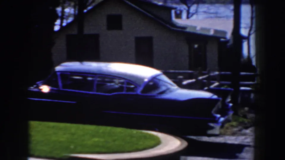 A finned two-tone car with a bright rear light is seen through a dark foreground, with lawn, driveway, houses, trees, and water behind it.