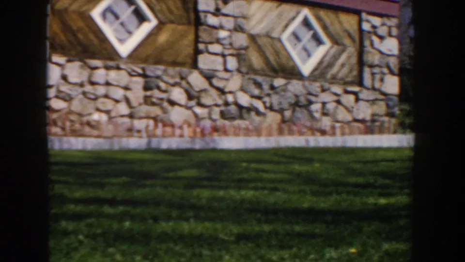 Panning 1959 Michigan frame with rough stonework, diagonal wood panels, two white diamond windows, and grass in front.