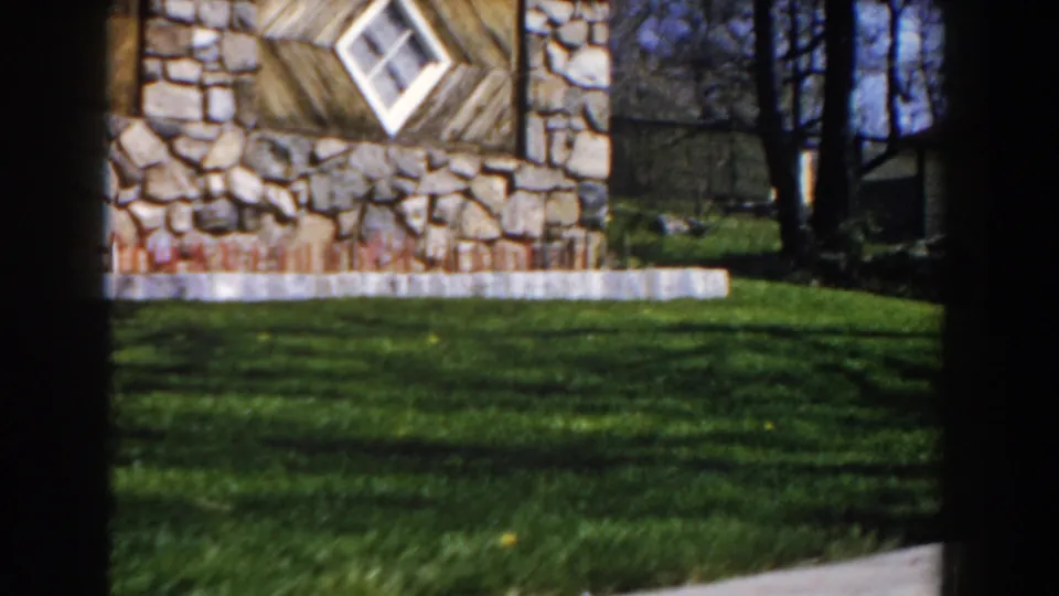 1959 Michigan home-movie frame showing the building corner, diamond window, green lawn, trees, and low concrete edging.
