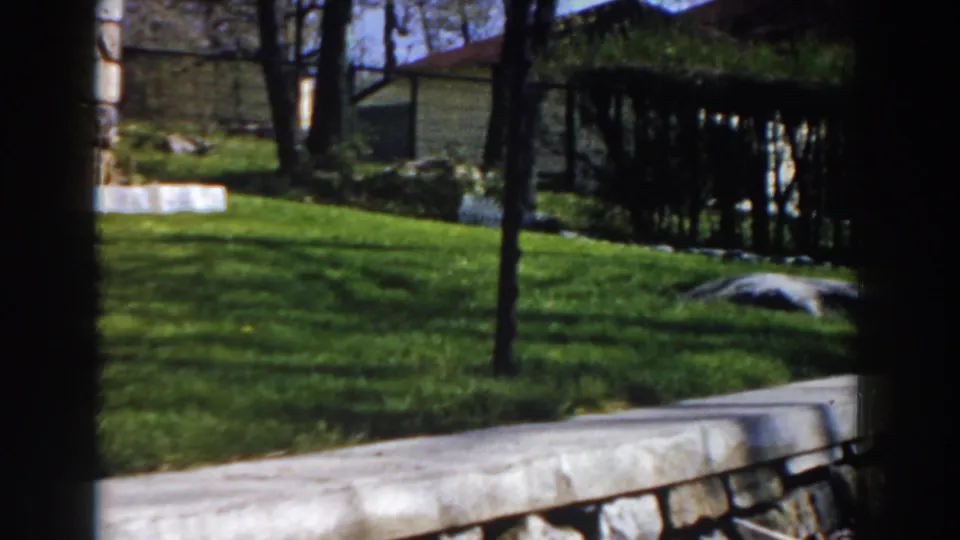 Late frame in the 1959 Michigan pan showing the lawn, trees, low retaining edge, and a partial rustic structure beyond.