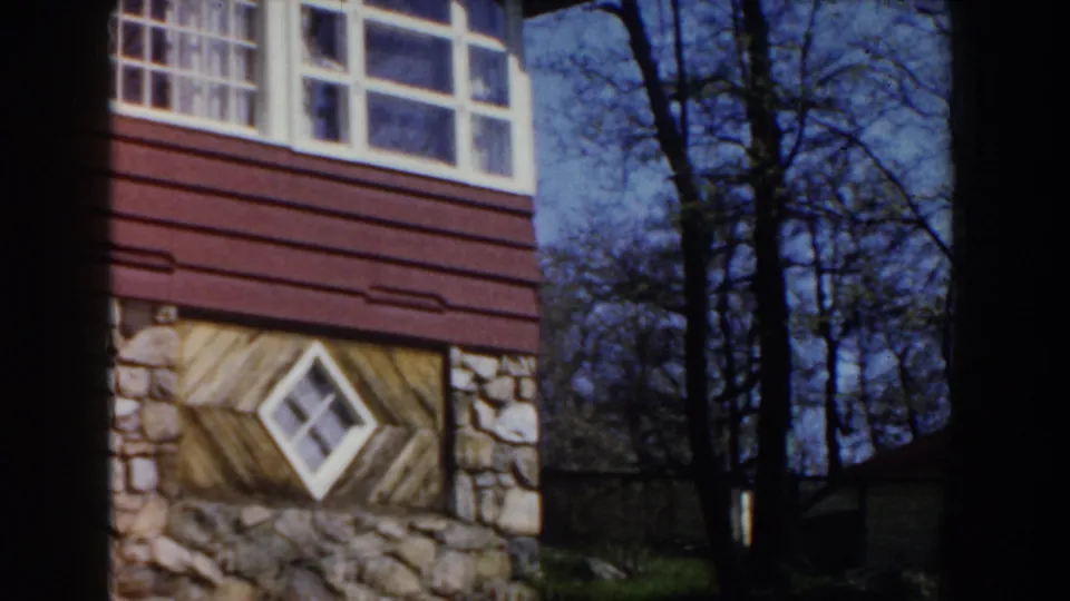1959 Michigan 8mm frame showing the building corner with stonework, red-brown siding, a diamond window, bare trees, and grassy ground.