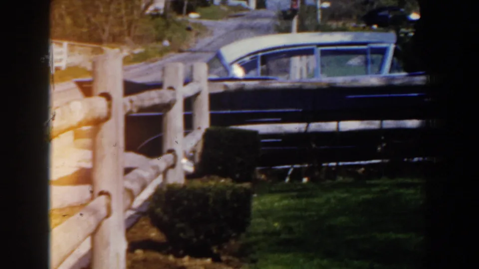 1959 Michigan frame showing a dark classic car parked by a weathered wooden fence, with grass, road edge, and bare trees nearby.